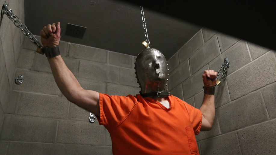 A prisoner gets chained in a jail cell wearing a metal head cage
