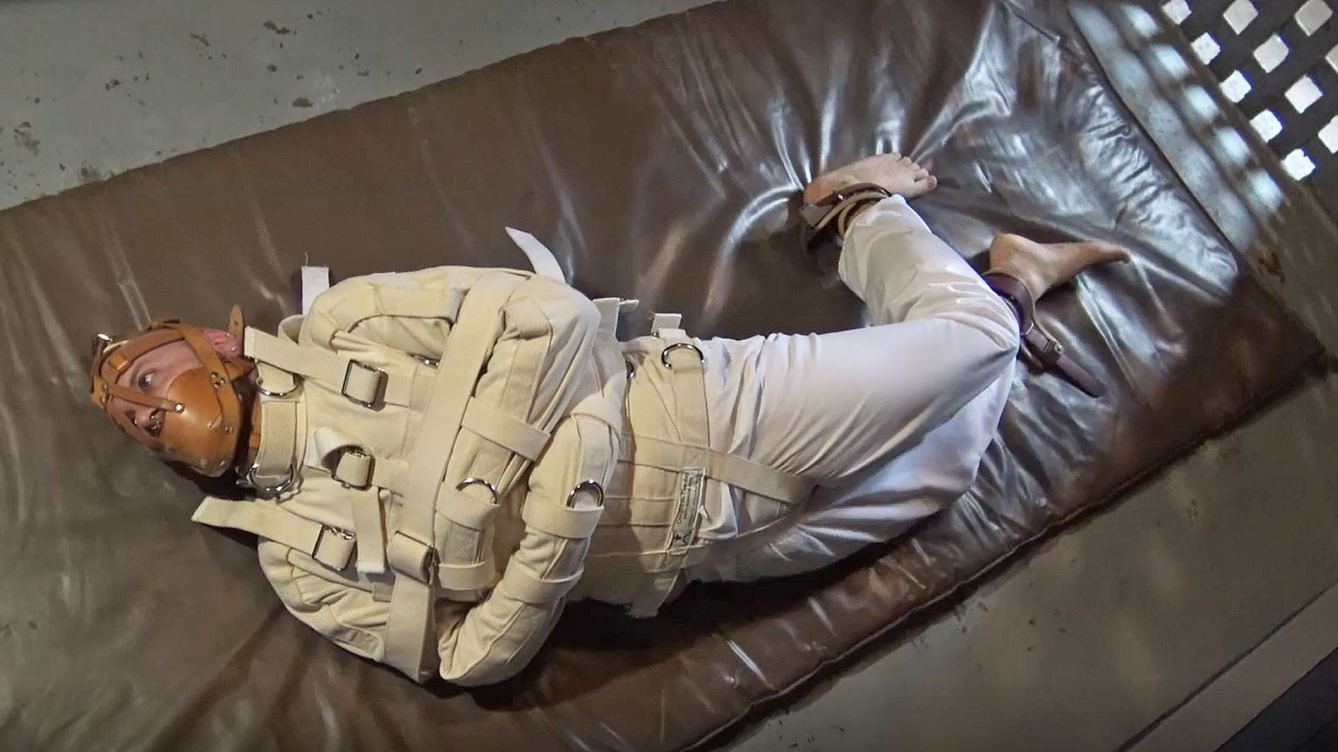 male captive bound in straitjacket inside a jail cell hogtied on a mat