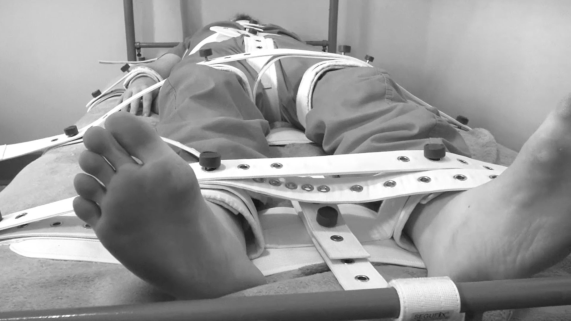 man strapped down in heavy bondage with bare feet visible