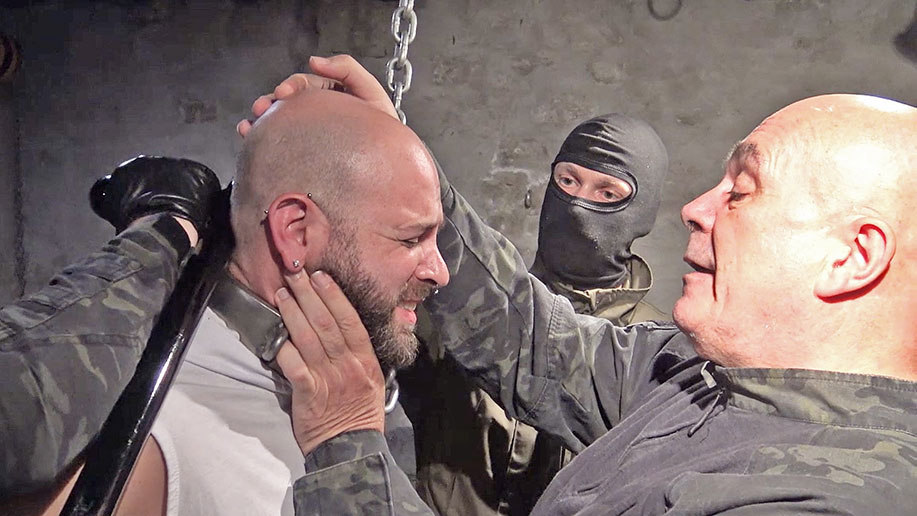a muscular man with a shaved head is chained up by sadistic prison guards
